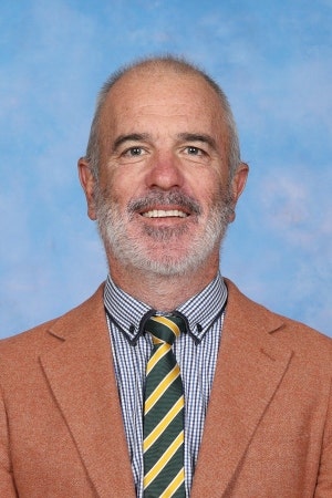 Portrait of the school principal smiling against a blue background, wearing a brown blazer, checkered shirt, and yellow-and-black striped tie.